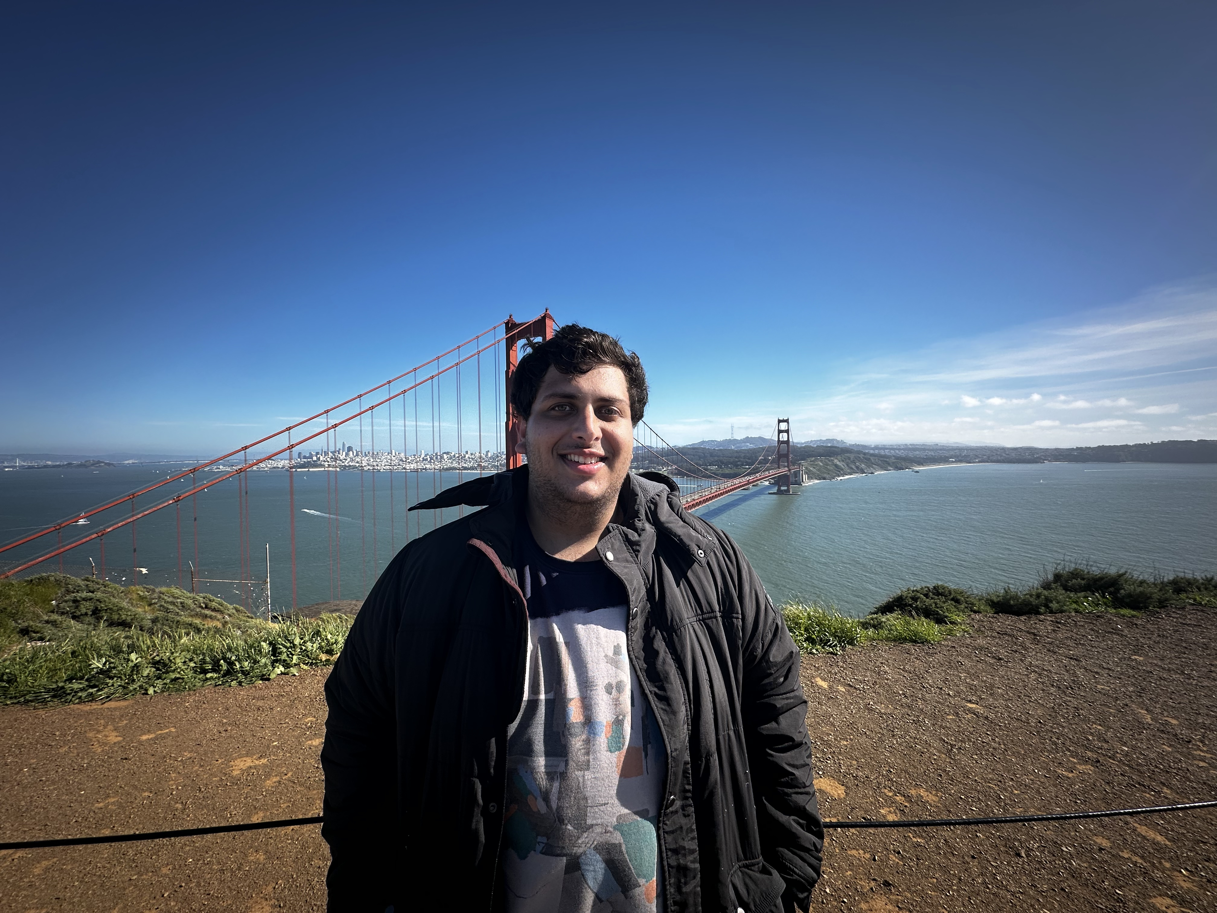 Ramneet standing in front of the Golden Gate Bridge wearing a blue t-shirt and a black jacket.